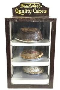 M1173 Fantastic very early 20th century Country Store bakery counter display case with lithographed tin advertising �Freihofer�s Quality Cakes� �A cake for every taste POUND SPONGE FRUIT�. Original lithographed marquee remains intact on top. Tin construction with three glass panels surrounding the front and sides and a solid faux wood grained hinged door on the back opens to reveal three tin display shelves inside. Circa 1913 � 1920s.