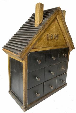 G525 Unique New England house-shaped nine drawer seed cabinet dated 1884 in original black and gold paint. The date applied to the front is made out of hand carved numbers. This date is also repeated in pencil on the back of the top row, center drawer, where the maker drew a �blueprint� of sorts when creating this project! A lot of details were put into the making of this piece, including lapped board shingles on the roof, double chimneys, nice moldings, original brass knobs and tapered outer corners on the top row�s outside drawers so as not to interfere with the roof line, or functionality of those drawers. It is all square head nail construction. Measurements: 16� wide at roof ends x 18 3/4� tall x 7 1/8� deep 