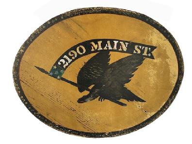 K1834 Double sided wooden sign with an Eagle holding a flag/banner in its beak that reads 2190 Main Street. Painted in black with polychrome accents on a mustard yellow colored background. Black border detail around the outside edges adds nice appeal. Old eye hooks remain intact on top and wire has been added for hanging purposes. Weathered surfaces indicate years of use in an outdoor setting. Measurements: 21 ½ wide x ¾ thick x 17 ¼ tall. 