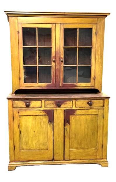 L469 Unique early 19th century Lancaster County, Pennsylvania original mustard painted two-piece stepback cupboard boasting an unusual configuration resting on an applied bracket base with cut out feet. The layout of the top consists of two fully mortised and pegged 6-windowlite doors that open to reveal shelves with plate grooves and a spoon rack along the front edge of the middle shelf. The layout of the lower part of the cupboard consists of three drawers over two mortised and pegged panel doors. The base of the cupboard is dovetailed. Nice, deeply beaded edges surround all doors and drawers and also adorn the internal edges of the backboards. The wood is pine with mortised/pegged with T nail and square head nail construction. Circa 1820s. Measurements: 57 ¼ wide (at waist) x 20 deep (at waist) x 87 tall.